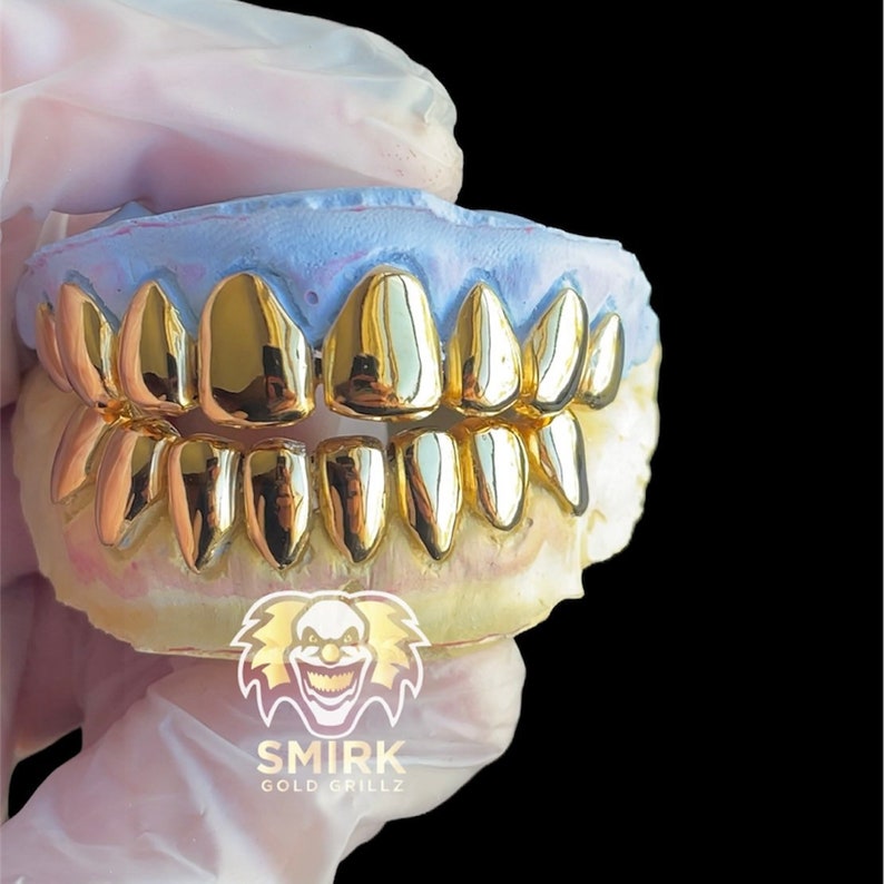 May include: Gold grillz with a shiny finish, displayed on a mold of a mouth. The grillz are made of multiple individual pieces that fit together to cover the front teeth. The mold is a light purple color. The image includes the text "SMIRK GOLD GRILLZ".