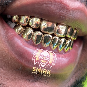 May include: Close-up of a person's mouth wearing a gold grill with a logo that says "Smirk Gold Grillz".