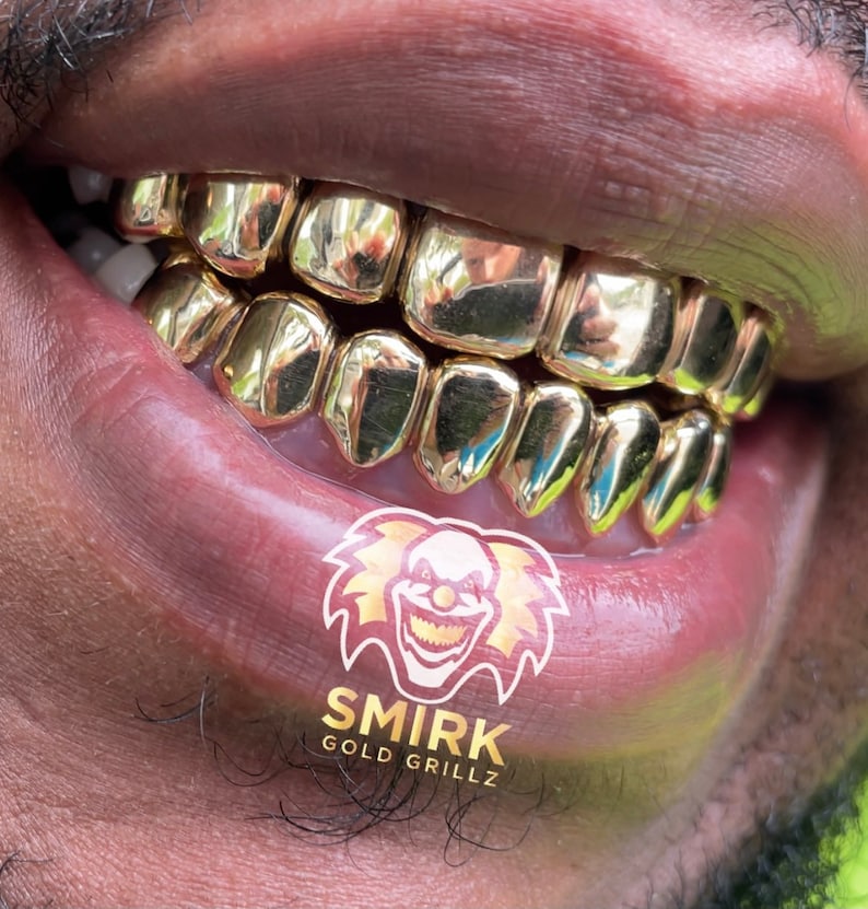 May include: Close-up of a person's mouth wearing a gold grill with a logo that says "Smirk Gold Grillz".