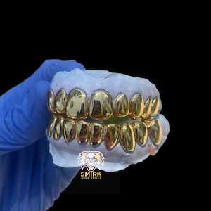 May include: Gold teeth grillz in a mold, with the brand name "Smirk Gold Grillz" printed below.