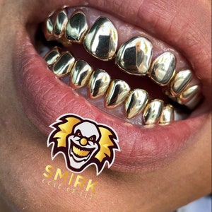 May include: Close-up of a person's mouth wearing gold grillz. The grillz are made of multiple individual pieces that fit over the teeth. The grillz are shiny and reflect light. The person's lips are closed and the grillz are visible. The image is cropped to show only the mouth and grillz.