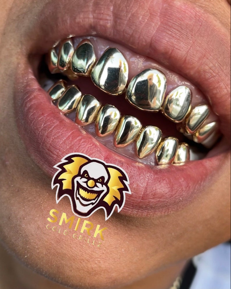 May include: Close-up of a person's mouth wearing gold grillz. The grillz are made of multiple individual pieces that fit over the teeth. The grillz are shiny and reflect light. The person's lips are closed and the grillz are visible. The image is cropped to show only the mouth and grillz.