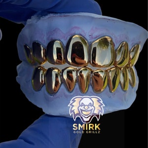 May include: Gold teeth grillz in a mold. The grillz are made of gold and have a shiny finish. The mold is blue and has a pink stain. The text "SMIRK GOLD GRILLZ" is visible at the bottom of the image.