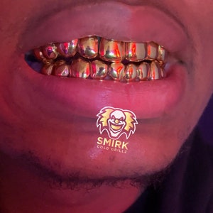 May include: Close-up of a person's mouth wearing gold grillz. The grillz are made of multiple individual pieces of gold that are connected together. The grillz are covering the person's teeth. The person's lips are closed and the grillz are visible. The image also includes a logo for "Smirk Gold Grillz" with a cartoon clown face.