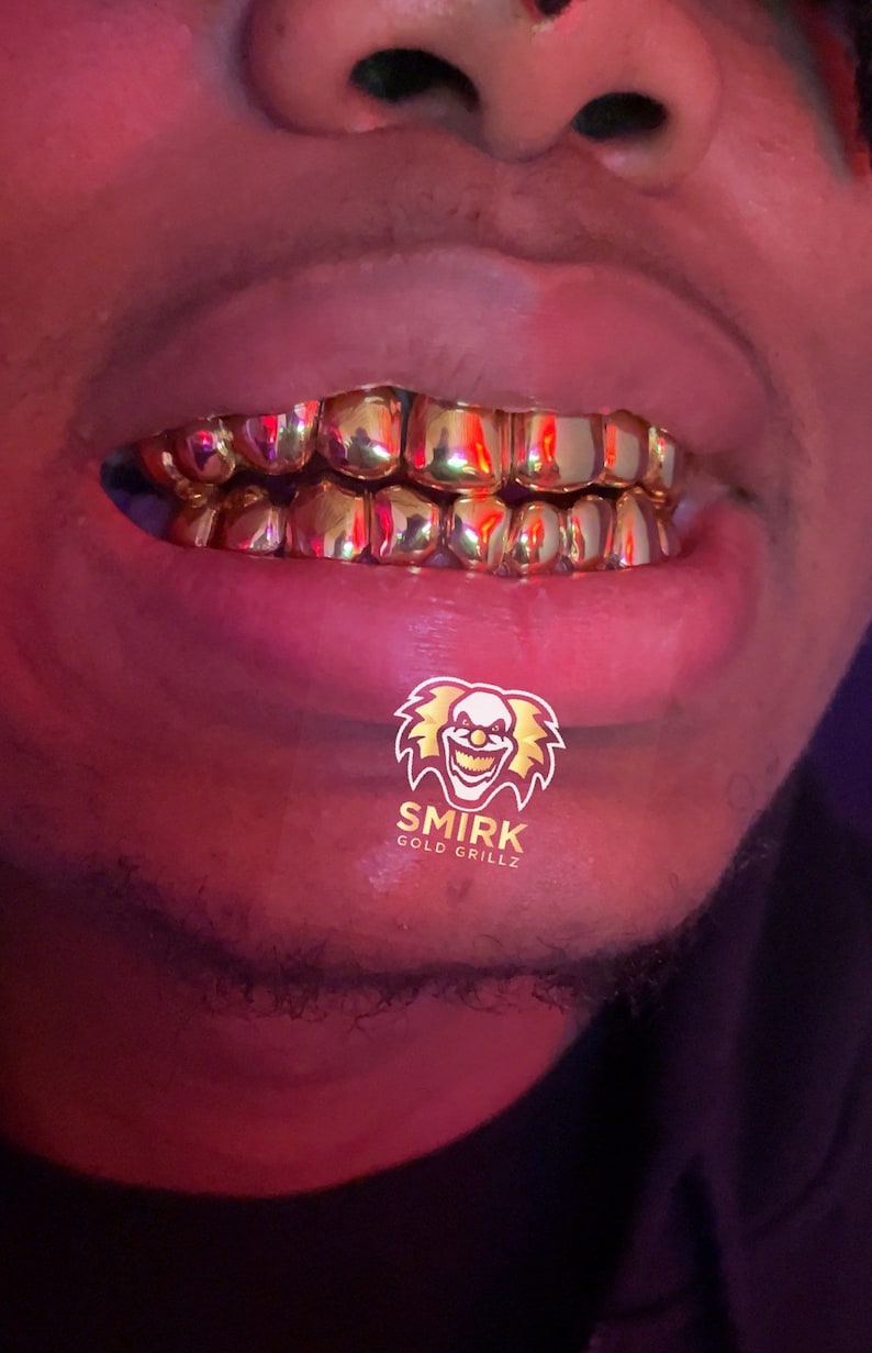 May include: Close-up of a person's mouth wearing gold grillz. The grillz are made of multiple individual pieces of gold that are connected together. The grillz are covering the person's teeth. The person's lips are closed and the grillz are visible. The image also includes a logo for "Smirk Gold Grillz" with a cartoon clown face.
