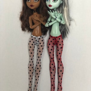 May include: Two fashion dolls with long hair wearing black and white polka dot tights and red and black polka dot tights. The doll on the left has brown skin and brown hair. The doll on the right has green skin and white and black hair.