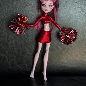 May include: A doll dressed in a red cheerleader outfit, holding two red and silver pom-poms. The doll has pink and black hair, pale skin, and a heart-shaped mark on its cheek. The outfit includes a crop top and skirt.