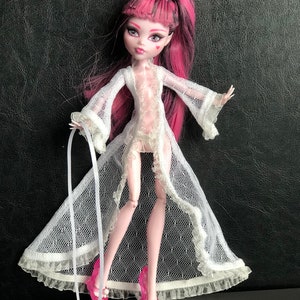 Pu&ograve; includere: Una bambola con i capelli rosa che indossa una vestaglia di pizzo bianco e tacchi alti rosa. La bambola ha un segno a forma di cuore sulla guancia.