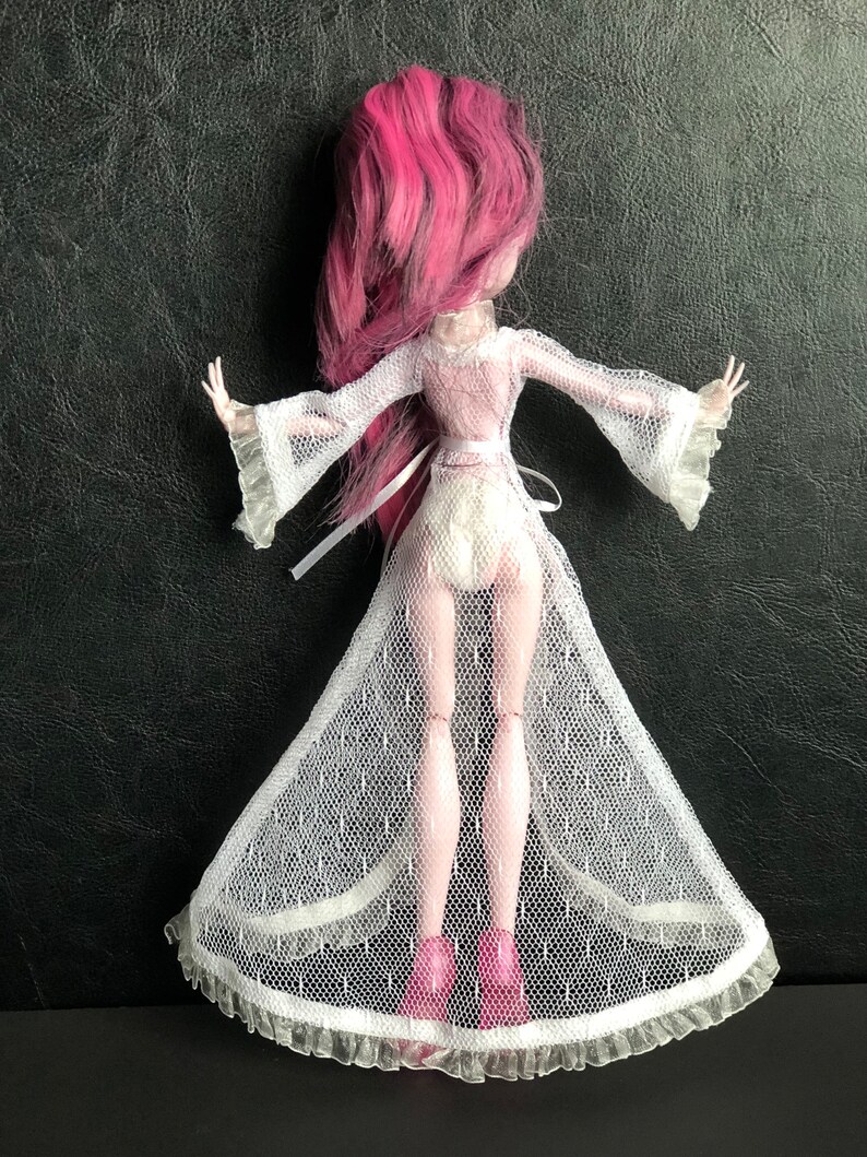 Pu&ograve; includere: Un abito da bambola in rete bianca con un nastro bianco e un bordo in pizzo. L'abito &egrave; indossato da una bambola con i capelli rosa.
