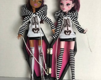 May include: Two fashion dolls with black and white striped skeleton print hoodies and matching striped knee-high socks. The dolls are standing in front of a pink and black coffin.