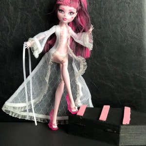 Pu&ograve; includere: Una bambola con capelli rosa e neri che indossa una vestaglia di pizzo bianca e tacchi alti rosa. La bambola &egrave; in piedi su una superficie nera con una striscia rosa.