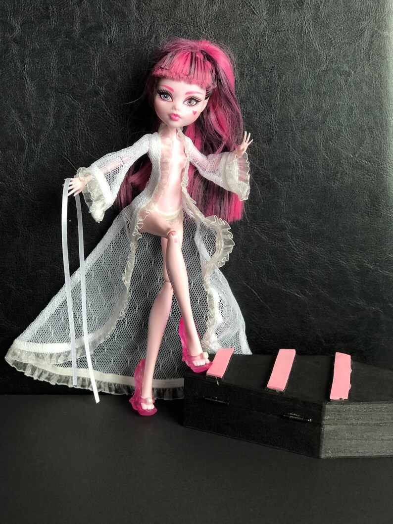 Pu&ograve; includere: Una bambola con capelli rosa e neri che indossa una vestaglia di pizzo bianca e tacchi alti rosa. La bambola &egrave; in piedi su una superficie nera con una striscia rosa.