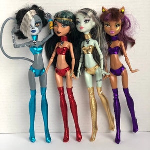 May include: Four Monster High dolls with different hairstyles and outfits. The dolls are wearing colorful, metallic thigh-high boots. The dolls are posed standing on a white surface.