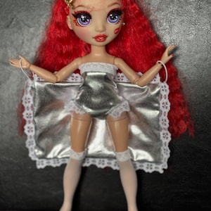 May include: A red-haired doll wearing a silver one-piece outfit with white lace trim and white stockings. The doll has bright blue eyes and red lipstick.