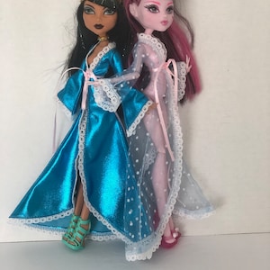 May include: Two fashion dolls wearing blue and pink satin robes with white lace trim. The doll on the left has dark brown skin, black hair, and a gold headband. The doll on the right has pink skin, pink hair, and a pink headband.