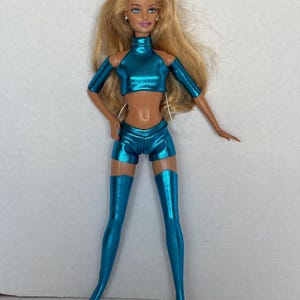 Peut inclure: Poupée mannequin blonde, vêtue d'un haut court métallique turquoise, d'un short et de cuissardes assortis. La poupée a le teint hâlé et est posée sur un fond uni. La tenue a un col montant et des manches découpées.