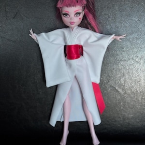 May include: A doll with pink hair and pale skin wearing a white kimono with a red obi. The doll has heart-shaped details on its face and is posed with arms outstretched.
