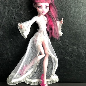 Pu&ograve; includere: Una bambola con capelli rosa e neri che indossa un abito bianco di pizzo e tacchi alti rosa. La bambola ha gli occhi azzurri e un trucco rosa.