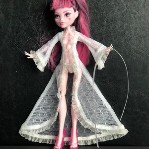 Pu&ograve; includere: Una bambola con capelli rosa e neri che indossa una vestaglia di pizzo bianca e tacchi alti rosa. La bambola ha un segno a forma di cuore sulla guancia.