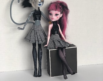 May include: Two fashion dolls wearing black and white plaid skirts, black tops, and black fishnet stockings. One doll has black and white hair and a cat tail. The other doll has pink hair and is sitting on a black cube.