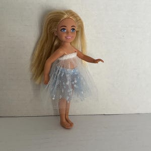 May include: A small doll with long blonde hair and blue eyes. The doll is wearing a white top and a light blue tulle skirt with white accents. The doll is standing on a white surface.