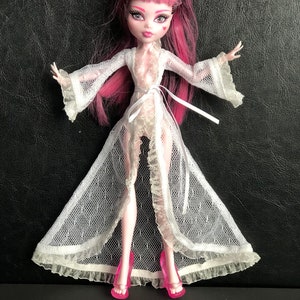 Pu&ograve; includere: Una vestaglia di pizzo bianco con un nastro rosa e tacchi alti rosa. La bambola indossa la vestaglia e ha i capelli rosa e neri.
