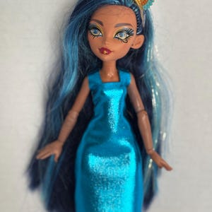 May include: A doll with long blue hair and a turquoise dress. The doll has a gold and turquoise headpiece. The doll's skin is tan, and it has detailed facial features, including blue eyes and red lips. The dress is a solid color with a square neckline.