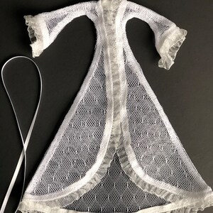 Pu&ograve; includere: Vestaglia di pizzo bianco con bordo a balze e gonna lunga e fluida. La vestaglia &egrave; aperta sul davanti e ha un laccio per legare.