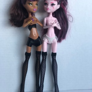 May include: Two fashion dolls with unique features. One doll has brown hair and cat ears, wearing a black top, shorts, and fishnet stockings. The other doll has pink skin, dark hair, and is wearing a white ruffled skirt and fishnet stockings.
