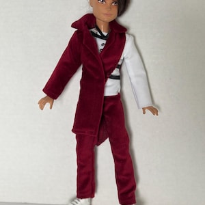 May include: A fashion doll in a burgundy velvet suit, comprising a long coat and trousers, paired with a white shirt. The doll has short, styled brown hair and white trainers. The outfit includes a black and white striped harness.