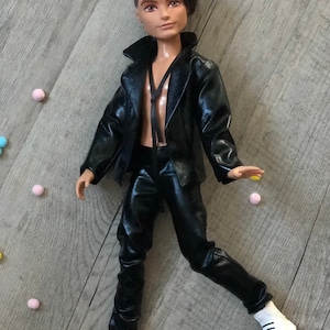 May include: A fashion doll with brown hair wearing a black leather jacket and trousers. The doll is wearing white trainers and has a black choker necklace.
