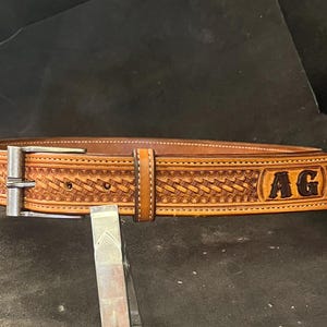 Peut inclure: Une ceinture en cuir marron avec un motif tissé et une boucle argentée. La ceinture porte les initiales "AG" en relief.
