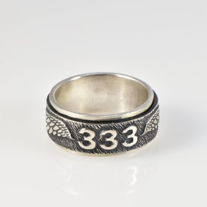 May include: A silver-toned metal ring featuring a textured, darkened band with the number 333 embossed in a lighter, polished finish. The band includes engraved wing-like details on either side of the numerals, set against a dark, brushed background.