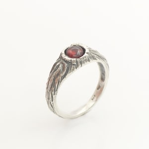 May include: A silver ring featuring a dark red, round, cabochon-cut gemstone set in a textured, bark-like metal band. The exterior of the band has deep, vertical grooves and a darkened finish, while the interior is smooth and polished silver.