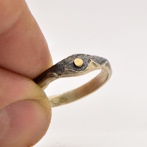 Könnte beinhalten: Ein Silberring mit einer dunklen, oxidierten und texturierten Oberfläche am oberen Band, in deren Mitte eine kleine, kreisförmige, goldfarbene Einlage glänzt. Das innere Band ist glatt und poliert, was dem Ring einen metallischen Glanz verleiht.