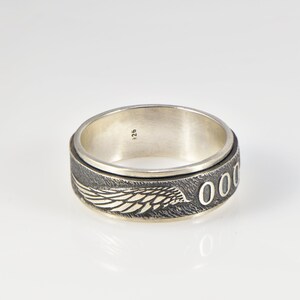 May include: A silver spinner ring featuring a dark, oxidized band with a textured surface. The band displays a detailed engraved wing design and a series of circular O shapes. The interior of the ring is smooth and stamped with the number 925.