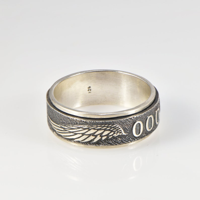 May include: A silver spinner ring featuring a dark, oxidized band with a textured surface. The band displays a detailed engraved wing design and a series of circular O shapes. The interior of the ring is smooth and stamped with the number 925.
