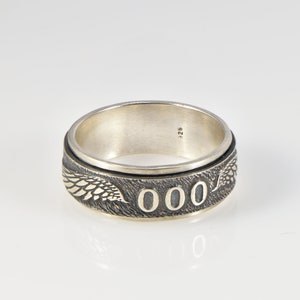 May include: A silver ring featuring a textured band with engraved wing motifs and the number 000 prominently displayed. The interior of the band is smooth and stamped with the hallmark 925, indicating sterling silver composition.