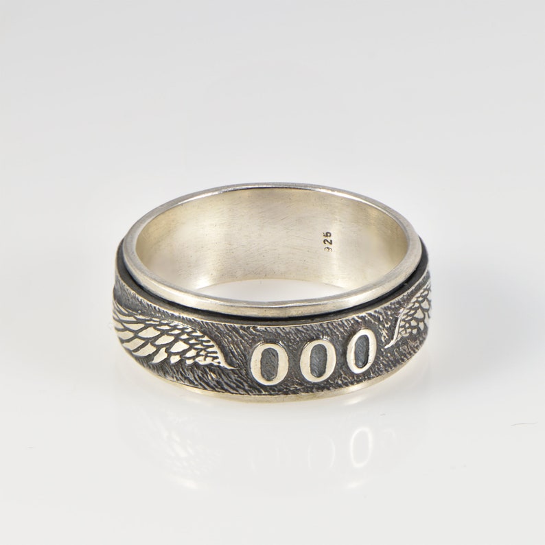 May include: A silver ring featuring a textured band with engraved wing motifs and the number 000 prominently displayed. The interior of the band is smooth and stamped with the hallmark 925, indicating sterling silver composition.