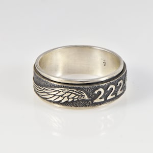 May include: A silver ring with a black and silver band. The band features a detailed wing design and the number "222" engraved in gold. The ring is marked with the size "SZ 25".