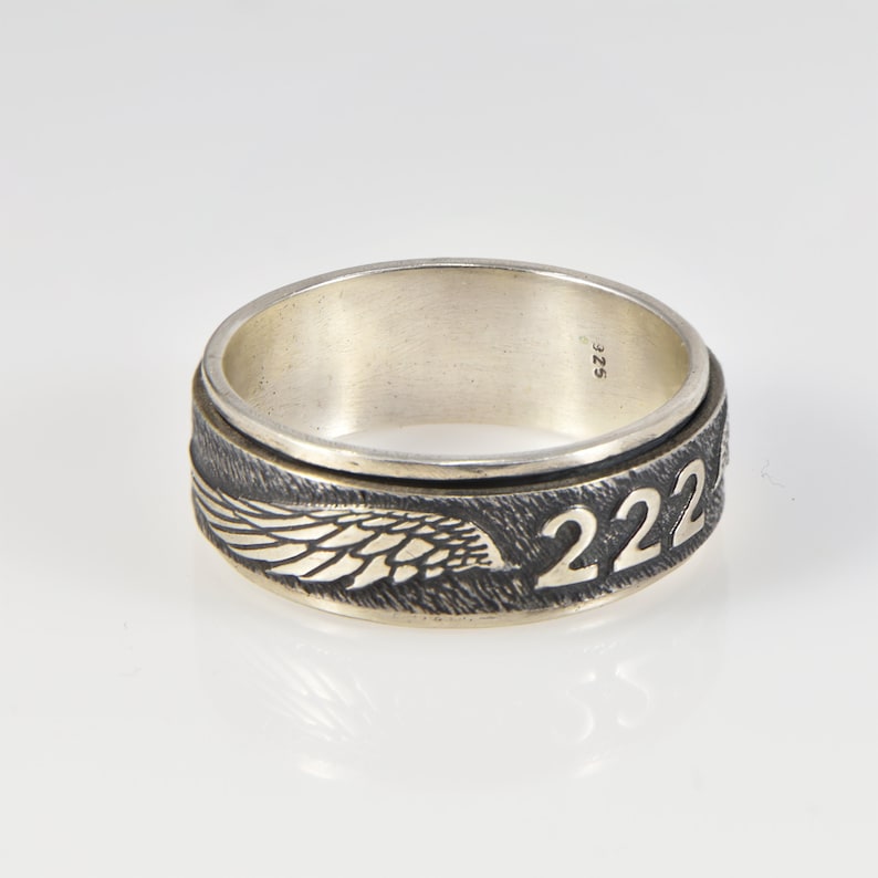 May include: A silver ring with a black and silver band. The band features a detailed wing design and the number "222" engraved in gold. The ring is marked with the size "SZ 25".