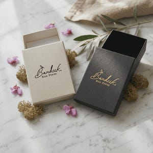 May include: Two rectangular sliding gift boxes, one in cream and one in black, each featuring the gold-colored logo Banduk Roe Press printed on the top surface. The boxes are open, revealing their interior structure and sturdy paper construction.