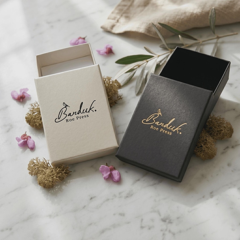 May include: Two rectangular sliding gift boxes, one in cream and one in black, each featuring the gold-colored logo Banduk Roe Press printed on the top surface. The boxes are open, revealing their interior structure and sturdy paper construction.