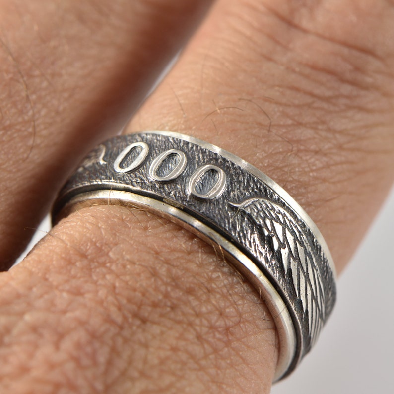 May include: A silver-toned metal ring featuring a textured, oxidized finish with the number 000 engraved on the surface. The band displays detailed wing-like patterns flanking the numbers, creating a distinct visual contrast against the dark background.