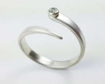 May include: A silver-toned bypass ring featuring a matte finish and an open design. One end of the band terminates in a tapered point, while the other end is capped with a single, small, round, clear faceted stone set in a smooth bezel setting.