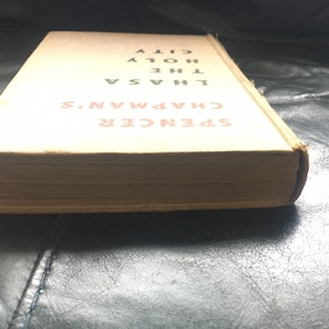 Puede incluir: Un libro antiguo titulado "SPENCER CHAPMAN'S LHASA THE HOLY CITY" con una cubierta de color beige y letras rojas.