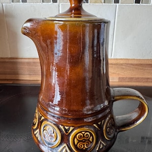 May include: A brown glazed ceramic pitcher with a matching lid, featuring a glossy finish and a textured band around the base decorated with repeating circular motifs and geometric shapes. The pitcher has a curved handle and a prominent pouring spout.