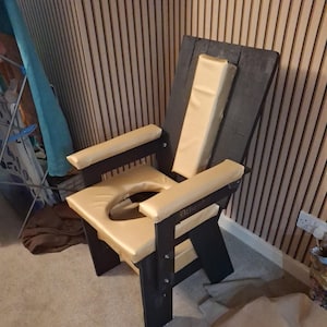 May include: A black wooden chair with a rectangular back and armrests. The seat and armrests are padded with a light tan material. The chair has a circular opening in the seat.