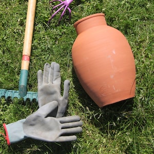 Puede incluir: Una maceta de terracota con base redonda y una abertura ancha. La maceta está sentada sobre un lecho de hierba verde. Hay dos guantes de trabajo grises y un rastrillo en la imagen.