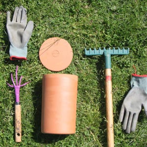 Pode incluir: Um conjunto de ferramentas de jardinagem que inclui um vaso de terracota, uma tampa de terracota, um ancinho com cabo de madeira, um ancinho de mão com cabo de madeira e dois pares de luvas de trabalho cinzas.